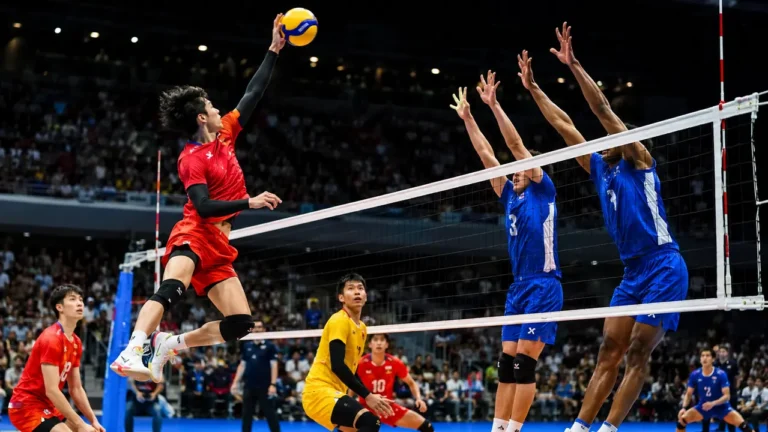Joueur de volley-ball sautant au filet lors d’un match intense en salle avec ballon, adversaires et couleurs vives.
