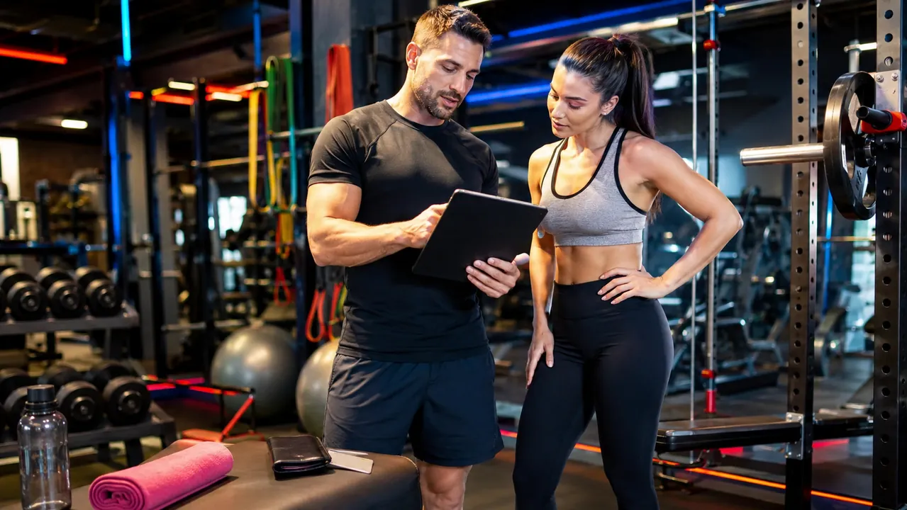 Coach sportif en salle accompagnant une cliente dans une salle de fitness moderne avec accessoires colorés et tablette.