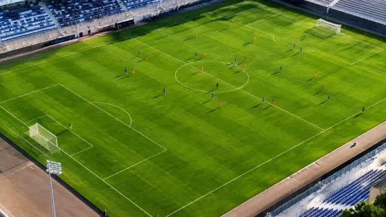 Vue aérienne d’un terrain de football officiel avec pelouse verte, lignes blanches réglementaires et joueurs répartis sur le terrain pendant un match.
